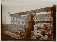 Photographie montrant deux mécaniciens nettoyant des pièces d'avion posées sur une caisse en bois devant l'appareil Zodiac 2S. Bâtiment et maison en arrière plan