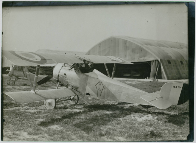 Photographie montrant un avion de chasse biplan français NIEUPORT 17 au sol, avec le pilote à son bord, devant des hangars