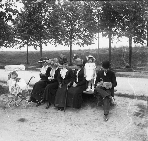 Groupe de cinq personnes assis sur un banc. Bébé dans un landau à côté du banc et jeune fille debout sur le banc