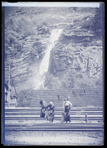 [Un homme et deux femmes traversant une voie ferrée à flanc de montagne. Au second plan : une cascade]