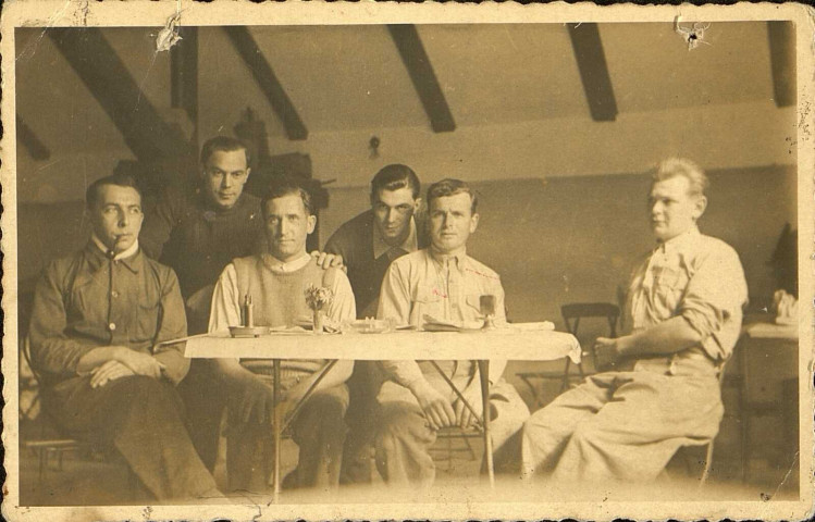 Prisonniers de guerre assis dans un baraquement du Stalag VIII-C. Louis Delignières est penché entre les deux prisonniers assis au centre