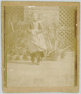 PHOTOGRAPHIE MONTRANT LE PORTRAIT, EN PIEDS, D'UNE JEUNE FILLE VETUE D'UNE ROBE TABLIER A CARREAUX. EN EXTERIEUR, DEVANT DES PLANTES VERTES EN POT. PALE. Lot de trente documents (26305 à 26334) provenant de la famille PAILLE-ROUGE de Ham. Damien PAILLE est tombé en 1916 à l'âge de vingt ans