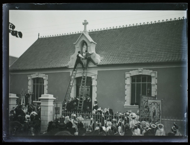 [Fête de Jeanne d'Arc à Camiers - octobre 1909. Devant la foule, deux hommes montés sur une échelle placent une statuette de la Vierge dans une niche sur la façade d'un édifice du village]