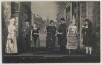Amiens. Scène d'une pièce de théâtre. Sept acteurs posent devant un décor représentant une rue. Une actrice arbore sur son costume une affiche "Maison à vendre présentement". Lucien Pilette se trouve au centre et porte un chapeau melon