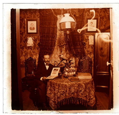 Intérieur d'une maison bourgeoise amiénoise. Un homme (Léon) assis consulte un album photographique de famille