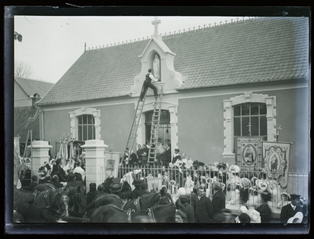 [Devant la foule, deux hommes montés sur une échelle placent une statuette de la Vierge dans une niche sur la façade d'un édifice du village]