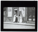 [Un homme sur un escabeau et deux femmes installant un drap blanc sur la façade d'une maison]