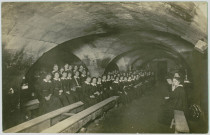 CARTE-PHOTO MONTRANT DES JEUNES FILLES PENSIONNAIRES ABRITEES DANS UNE CAVE. ASSISES EN RANG