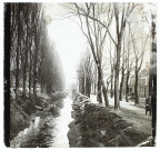 [Un cours d'eau bordé d'arbres aux abords de maisons d'habitation d'une ville. Dans la rue : un militaire et un homme sur un tricycle]