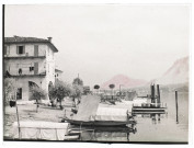 [Locarno : embarcations et maisons au bord du lac. Le photographe a légèrement rehaussé de couleur rouge les montagnes]