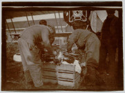 Photographie montrant deux mécaniciens réparant une pièce d'avion posée sur une caisse en bois devant l'appareil Zodiac 2S. Bâtiment et maison en arrière plan