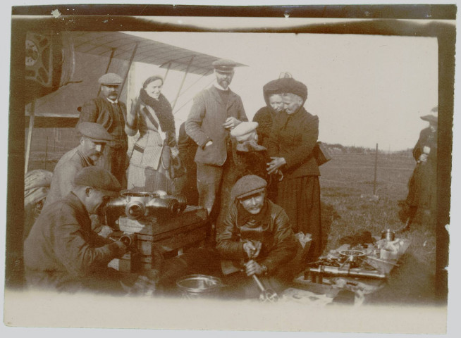 Photographie montrant des civils (hommes, femmes et un enfant) et des mécaniciens nettoyant des pièces posées sur des caisses en bois. Sur le côté gauche, un avion ZODIAC 2S