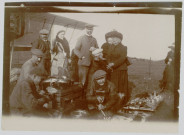 Photographie montrant des civils (hommes, femmes et un enfant) et des mécaniciens nettoyant des pièces posées sur des caisses en bois. Sur le côté gauche, un avion ZODIAC 2S