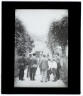 [Les membres de la Société Photographique de Picardie discutant dans une allée bordée d'arbres. Au fonds, vestige d'un portail]