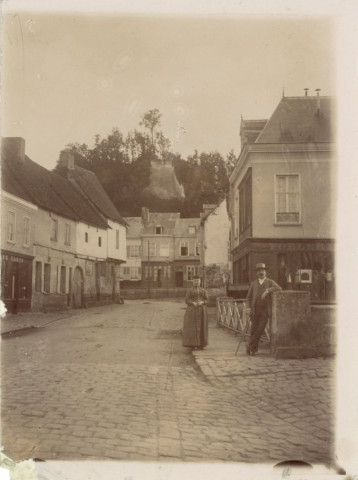 Picquigny. Photographie d'un homme et une femme sur un pont. Maisons et commerces dont une "poêlerie" et le "café Caron". Au second plan, sur les hauteurs, les ruines du château de Picquigny