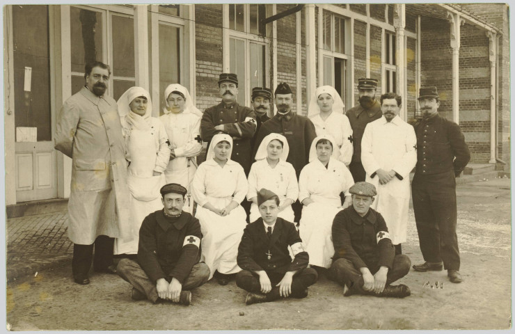 CARTE-PHOTO MONTRANT SEIZE PERSONNES ENGAGEES DANS LA CROIX-ROUGE ET POSANT POUR LA PHOTOGRAPHIE (OFFICIERS, MEDECINS, INFIRMIERES ET CIVILS) DANS UNE COUR. JAUNIE. TACHES JAUNATRES