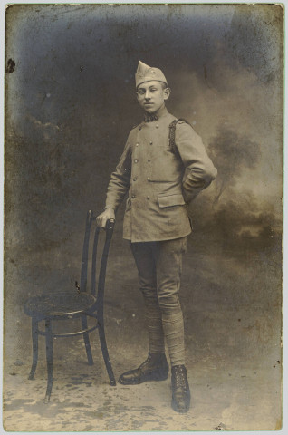 CARTE-PHOTO PRISE EN STUDIO MONTRANT LE PORTRAIT, EN PIEDS, D'UN SOLDAT FRANCAIS, APPUYE SUR UNE CHAISE