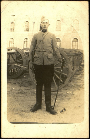 Photographie d'un jeune soldat dans la cour d'une caserne près de charriots de munitions