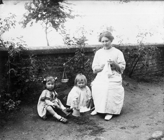 Femme assise brodant le long d'un mur avec deux jeunes filles assises et jouant à ses côtés